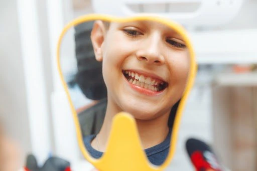 unnamed-11 A smiling child looks into a tooth-shaped mirror during a dental checkup, happily examining their clean teeth.